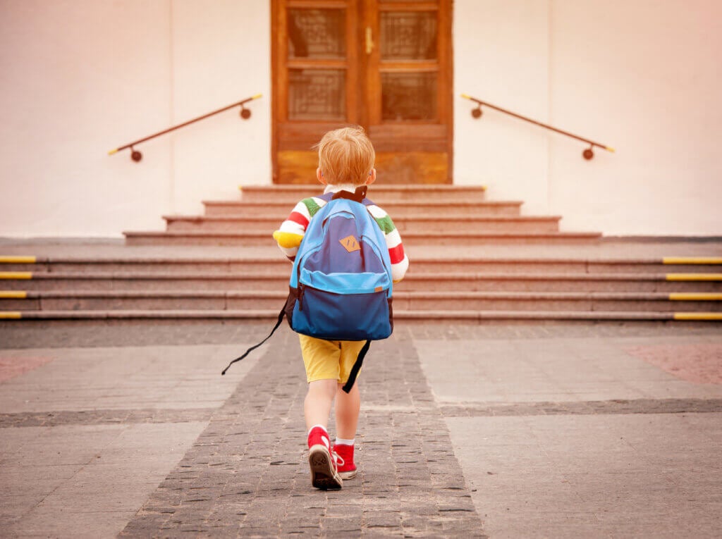 Little boy poses for back to school photo of him walking into school.