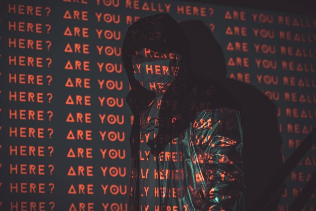 Person stands in dark with the phrase "Are you here?" repeatedly projected across them in red light.